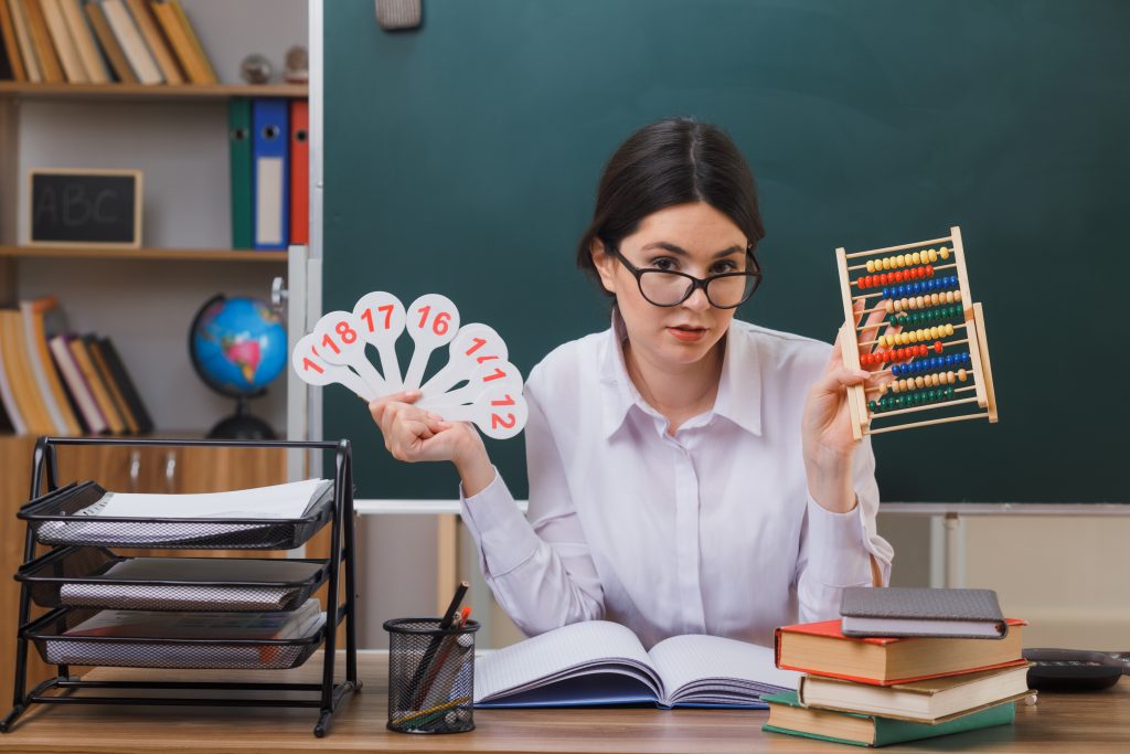 pleased young female teacher wearing glasses holding abacus with number fan sitting at desk with school tools in classroom