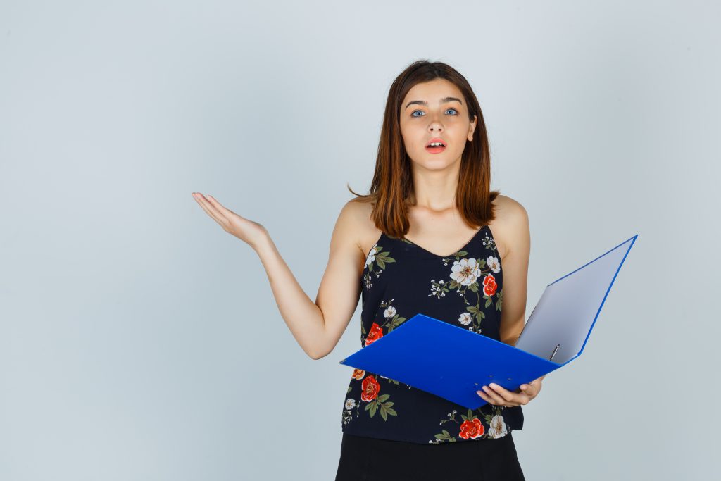 Portrait of young lady holding folder while showing helpless gesture in blouse, skirt and looking puzzled front view tnpsc study materials